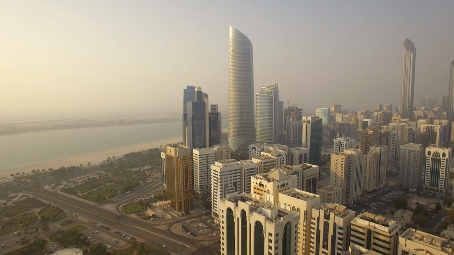 A hazy aerial view of Abu Dhabi’s skyline with modern high-rise towers, including a tall curved glass skyscraper at the center. The city’s grid of roads and beige residential buildings stretch outward, while the coastline with sandy beaches and calm waters appears to the left.